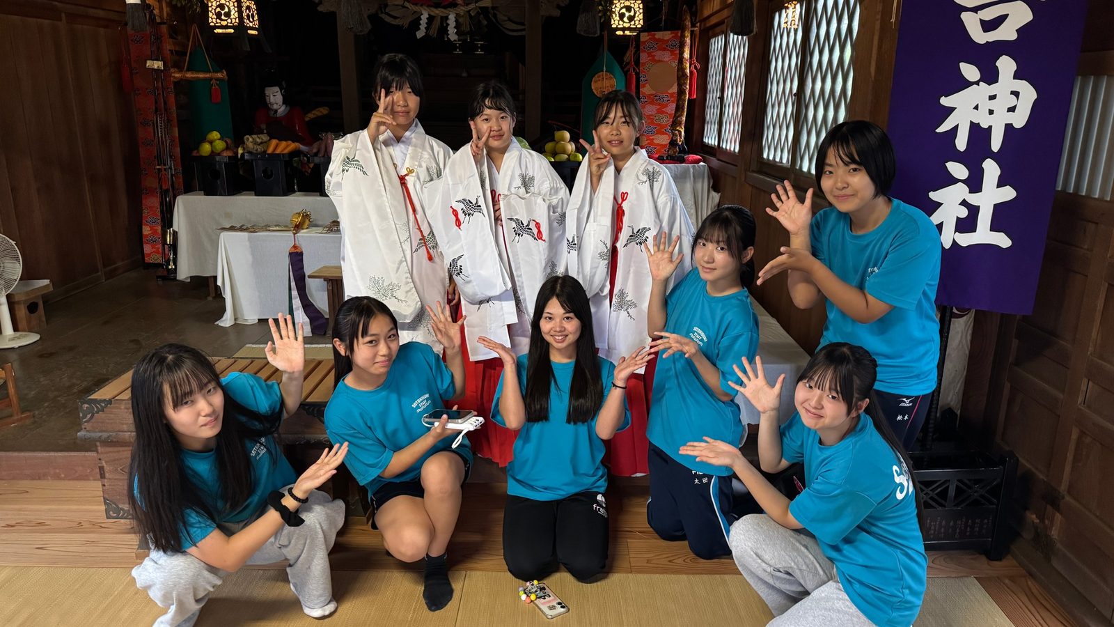 神社の拝殿で、装束を着たメンバーとTシャツ姿のメンバーが並ぶ様子